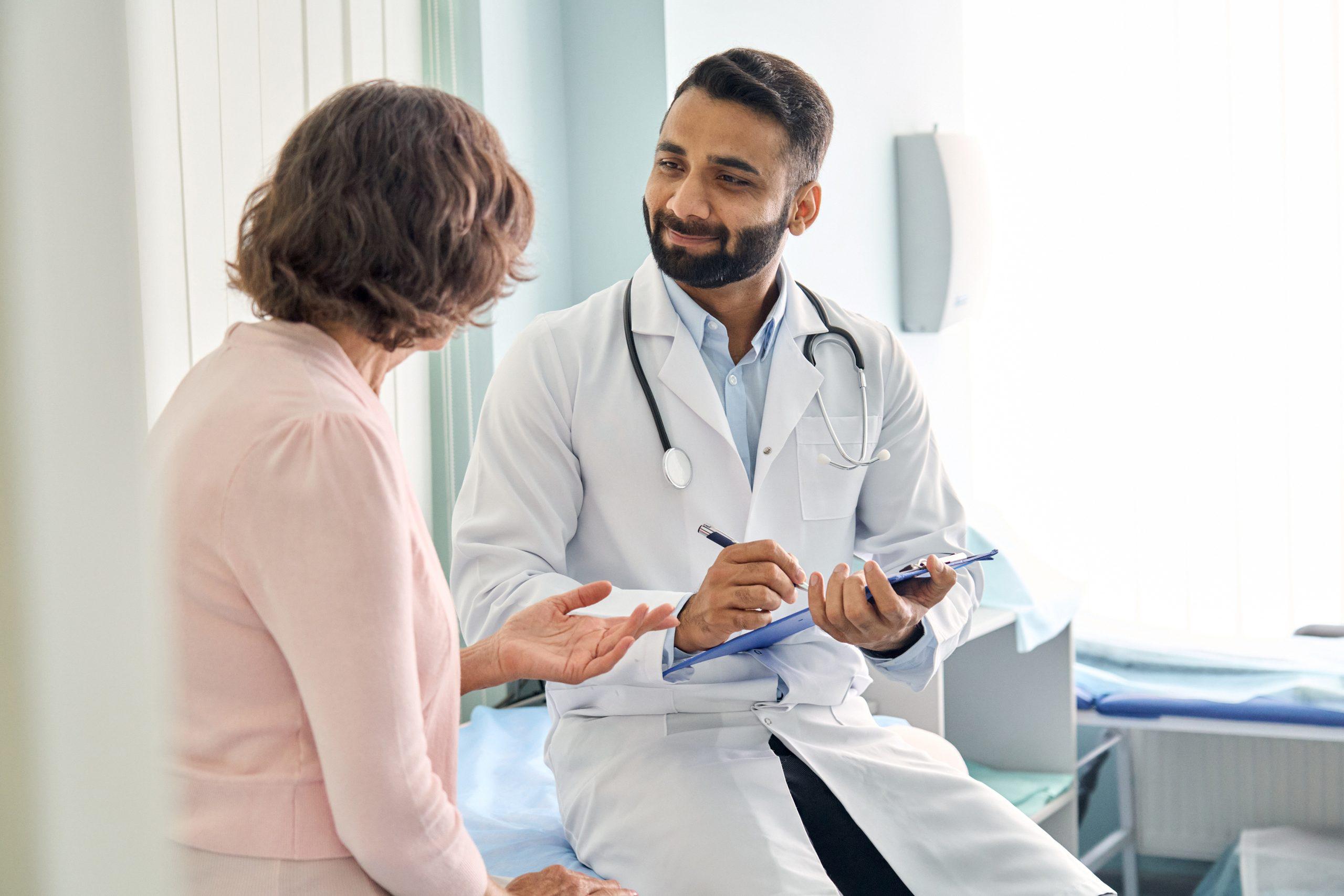  Indian male doctor consulting senior mature patient filling form at
consultation. Professional physician wearing white coat talking to mature woman signing
medical paper at appointment visit in clinic.