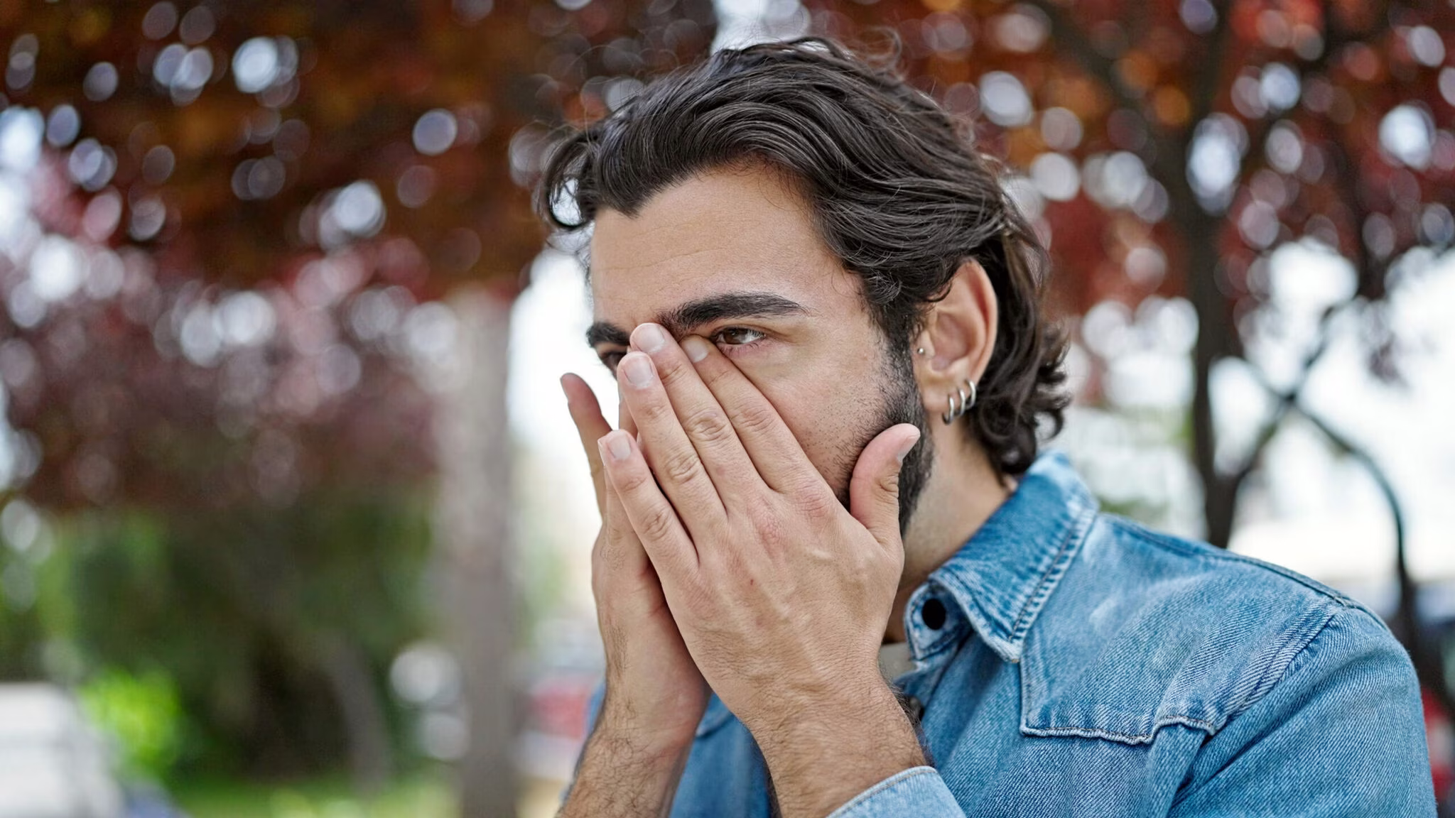 Young Hispanic Man rubbing his face due to allergies while walking in the park.