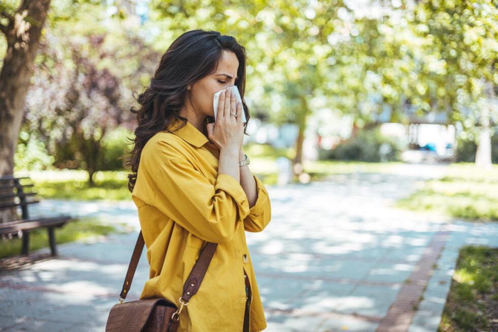 Woman in yellow shirt with brown crossbody purse slung over her shoulder, walking in park during spring, stopping to sneeze or blow her nose into facial tissue. Concrete pathway and bench in the background.