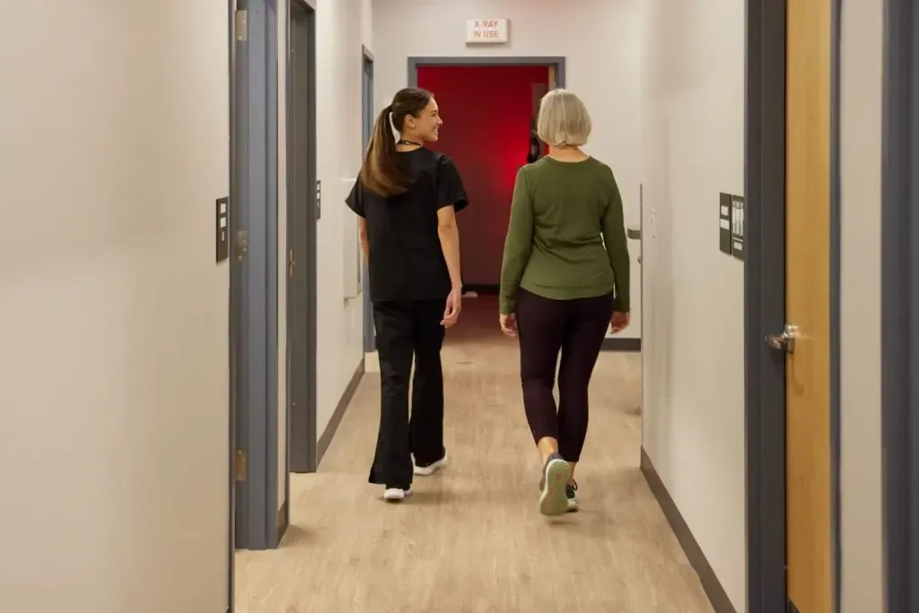 Two people walk down a narrow, well-lit hallway with blue doors. One wears scrubs and the other casual workout attire. The sign above reads "X RAY IN USE" in this bustling walk-in clinic where urgent care is provided, including physicals and other essential medical services.