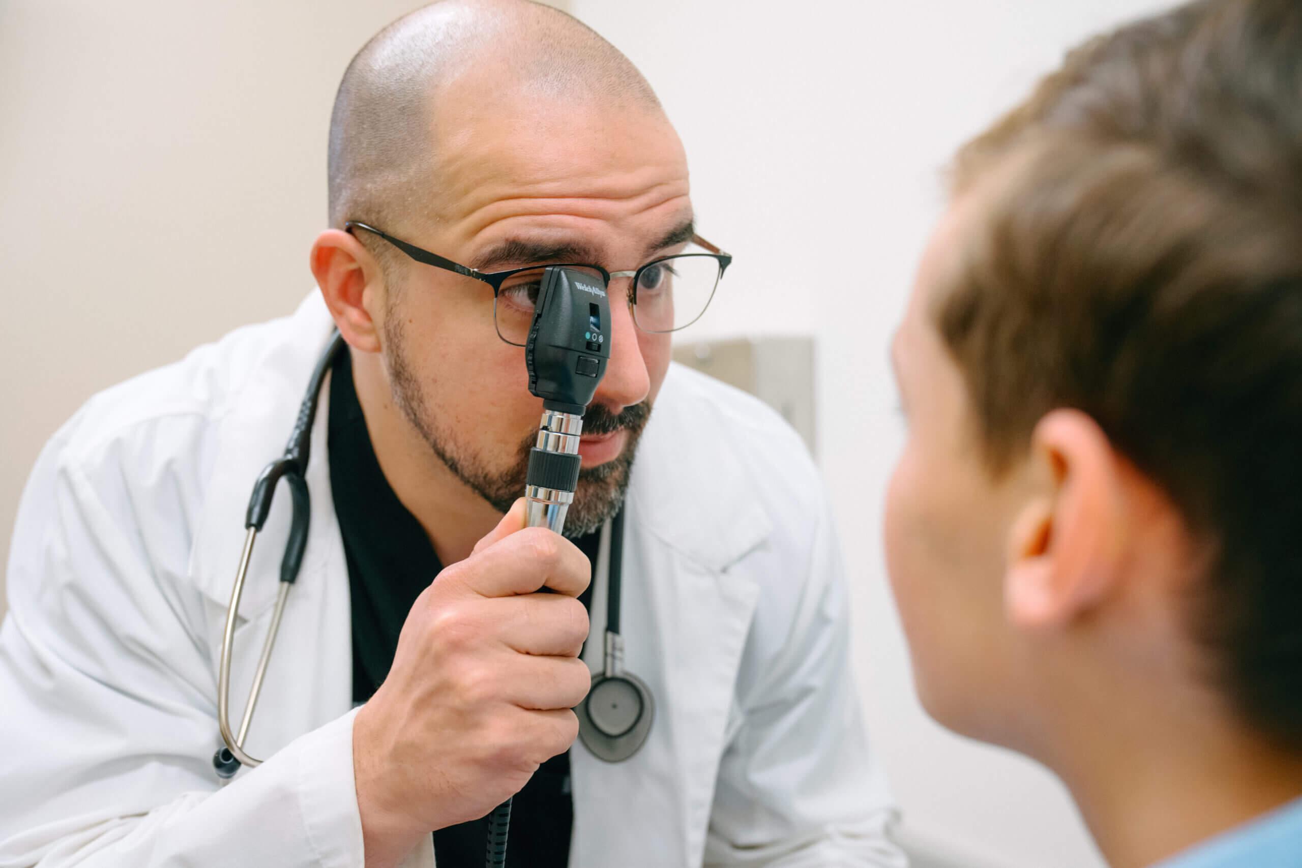 AFC Urgent Care provider performs a pink eye examination on a young male patient using an ophthalmoscope in an exam room. The provider wears a white coat and stethoscope while evaluating eye redness and conjunctivitis symptoms.