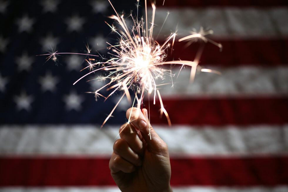 Close-up of a single right hand holding a lit sparkler in front of a softly waving American flag during a Fourth of July celebration.
