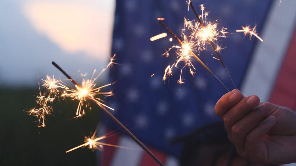 Close-up of people holding lit sparklers at dusk, with glowing sparks illuminating a right hand in the foreground and an American flag waving softly in the background during a Fourth of July celebration.