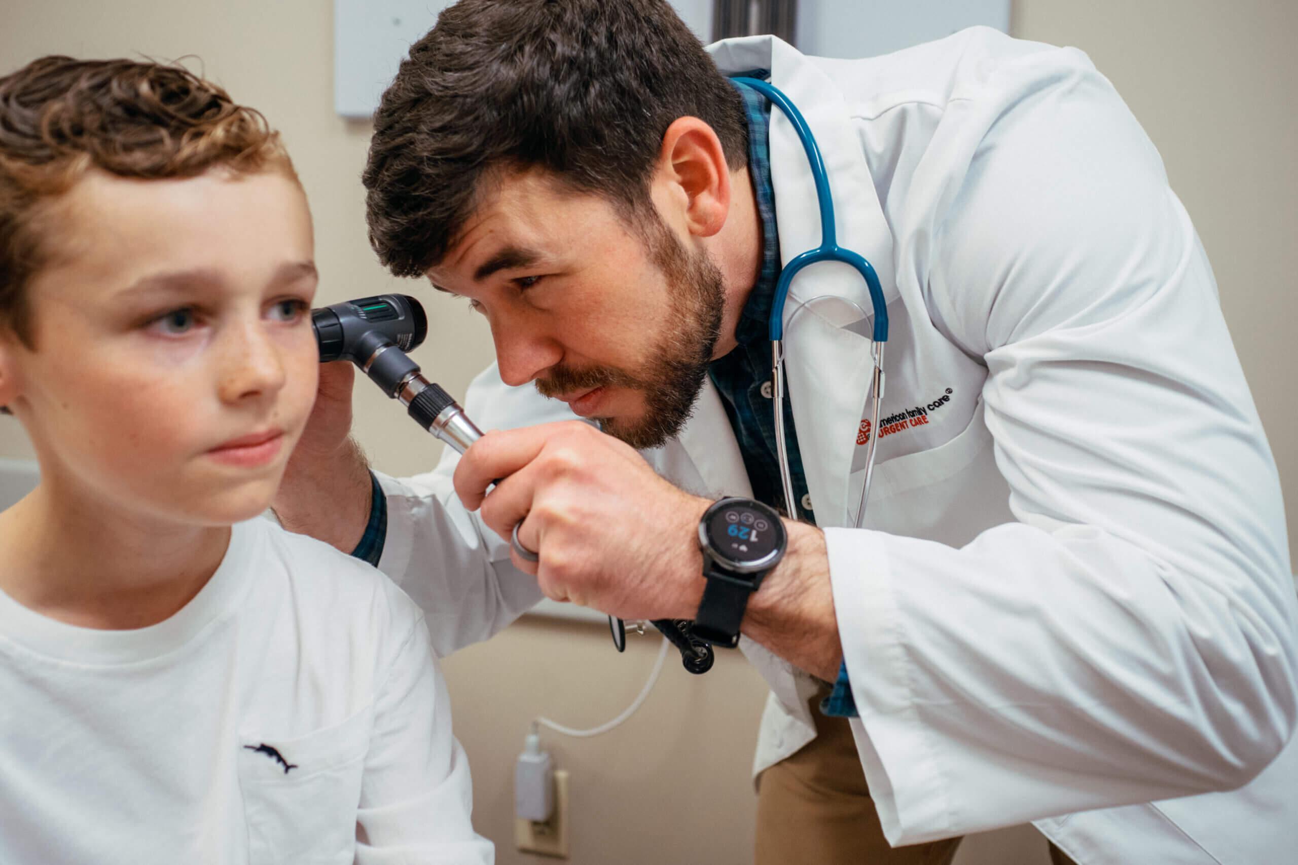 Male AFC Healthcare provider checking a pediatric patient's left ear with an otoscope during a pediatric physical examination.
