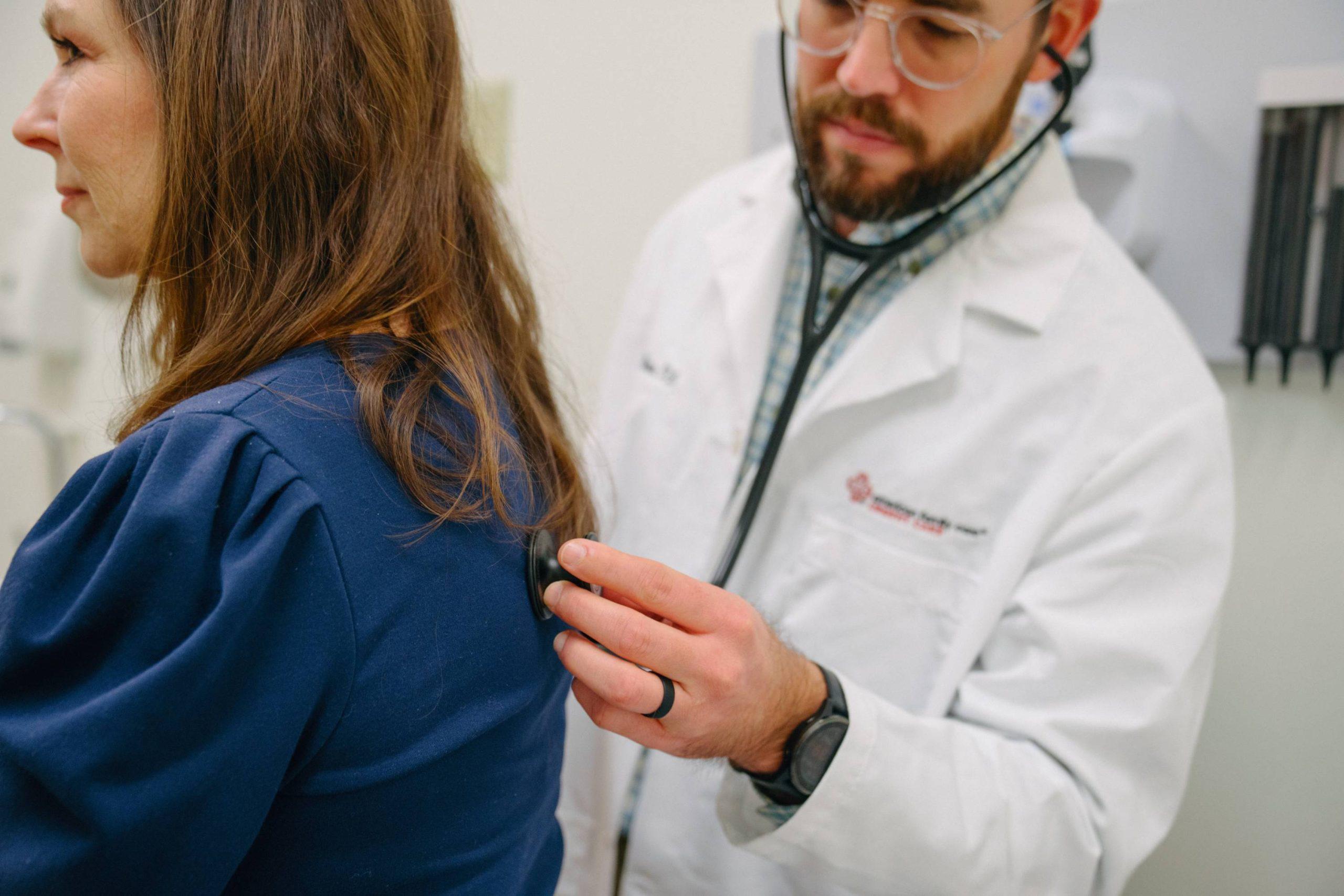  Close-up of an adult woman sitting on an exam table while an AFC healthcare provider listens to her breathing with a stethoscope during an evaluation for pneumonia symptoms at an urgent care clinic.