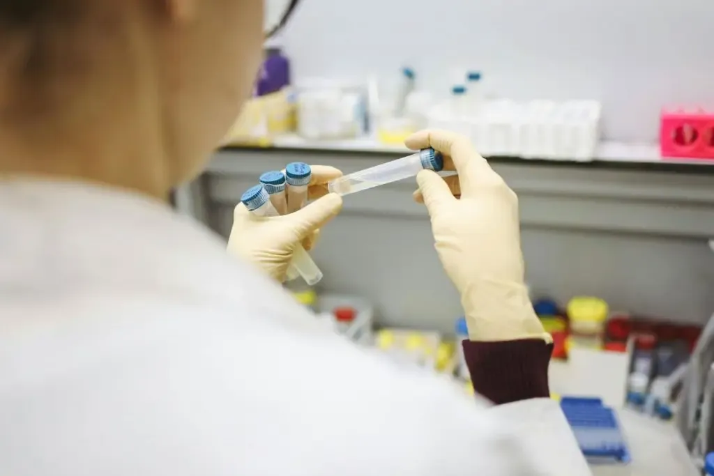 A person wearing gloves holds a test tube in one hand and a small vial in the other, in a medical center's laboratory with various lab equipment in the background.