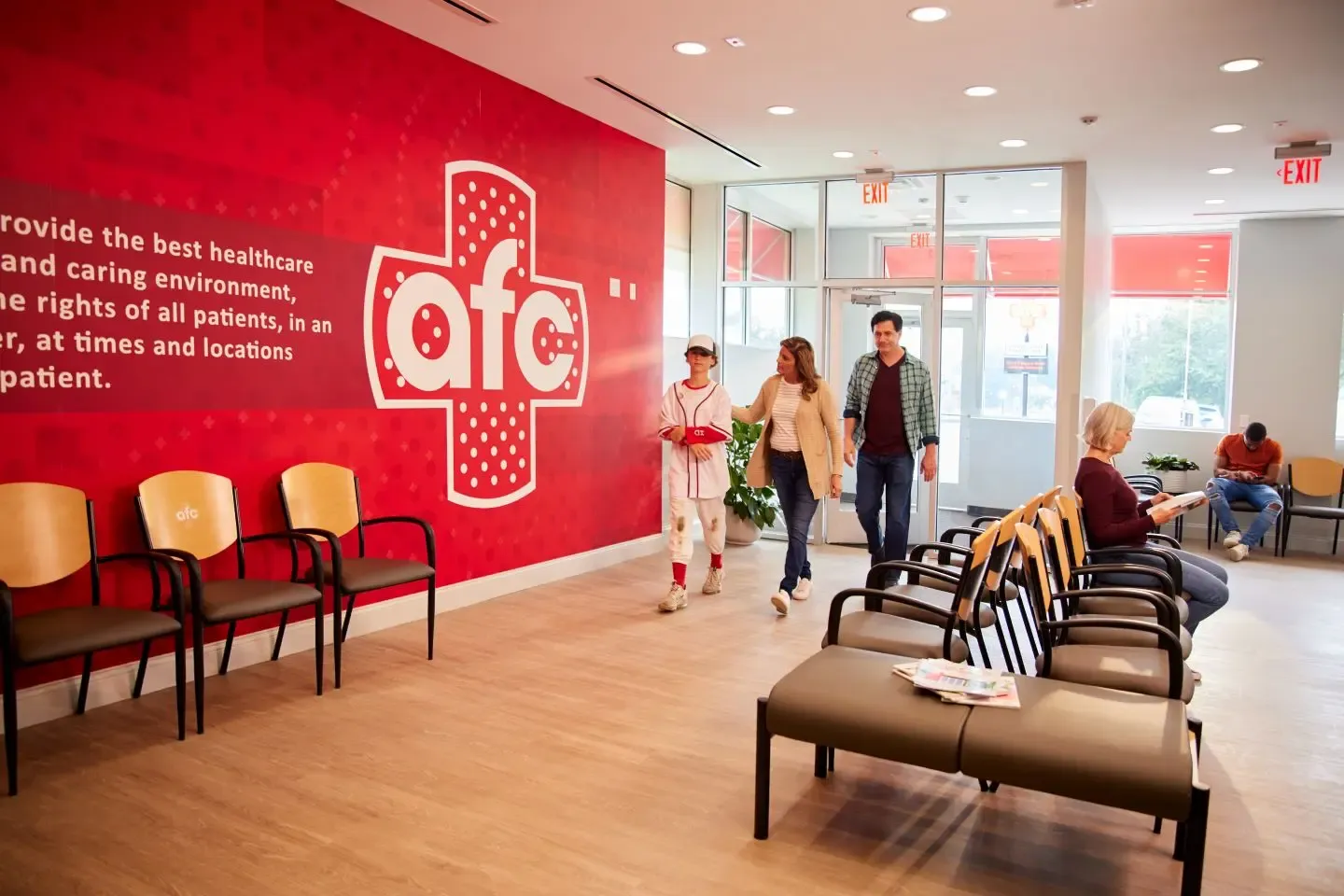 Waiting Area in a Healthcare Facility: The image depicts an interior space, likely a waiting area in a healthcare facility. Rows of chairs are arranged along the left wall, and there’s a red accent wall with partially visible white text. People are either seated or walking, and large windows allow natural light into the room.