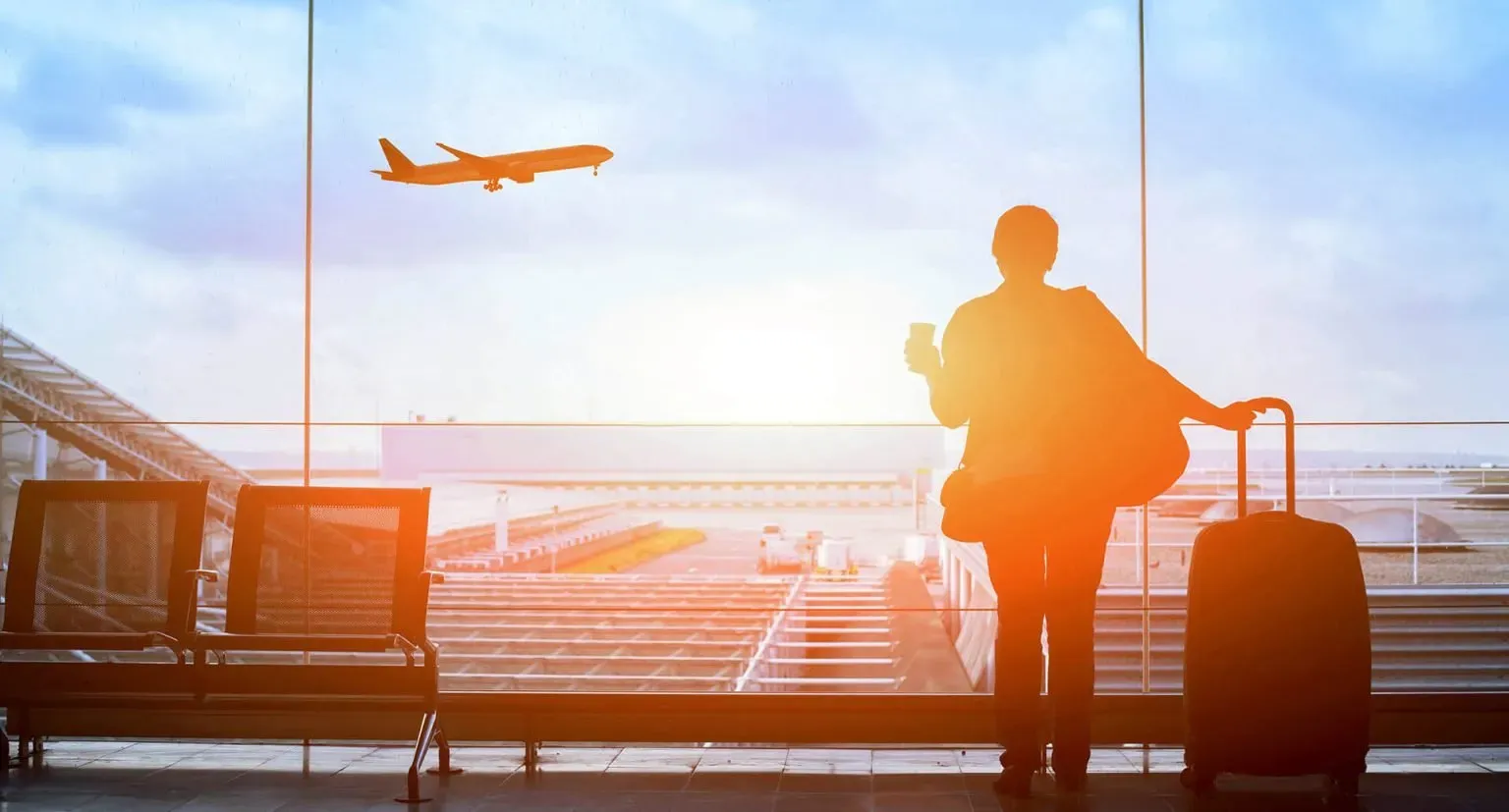 The back of a woman standing in the airport looking out the window at a plane taking off