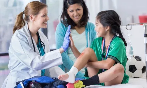 A doctor high-fives a young girl in a green sports uniform with a knee injury, while a woman, possibly her mother, smiles next to them in the medical center.