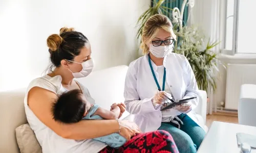 A mother holds her baby while sitting on a couch next to a healthcare professional in an urgent care center, both wearing masks. The healthcare professional is taking notes on a tablet.